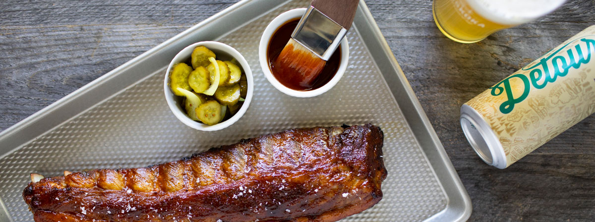 Rack of ribs, dish of sliced pickles, dish of bbq sauce with brush in it, can and glass of Detour.